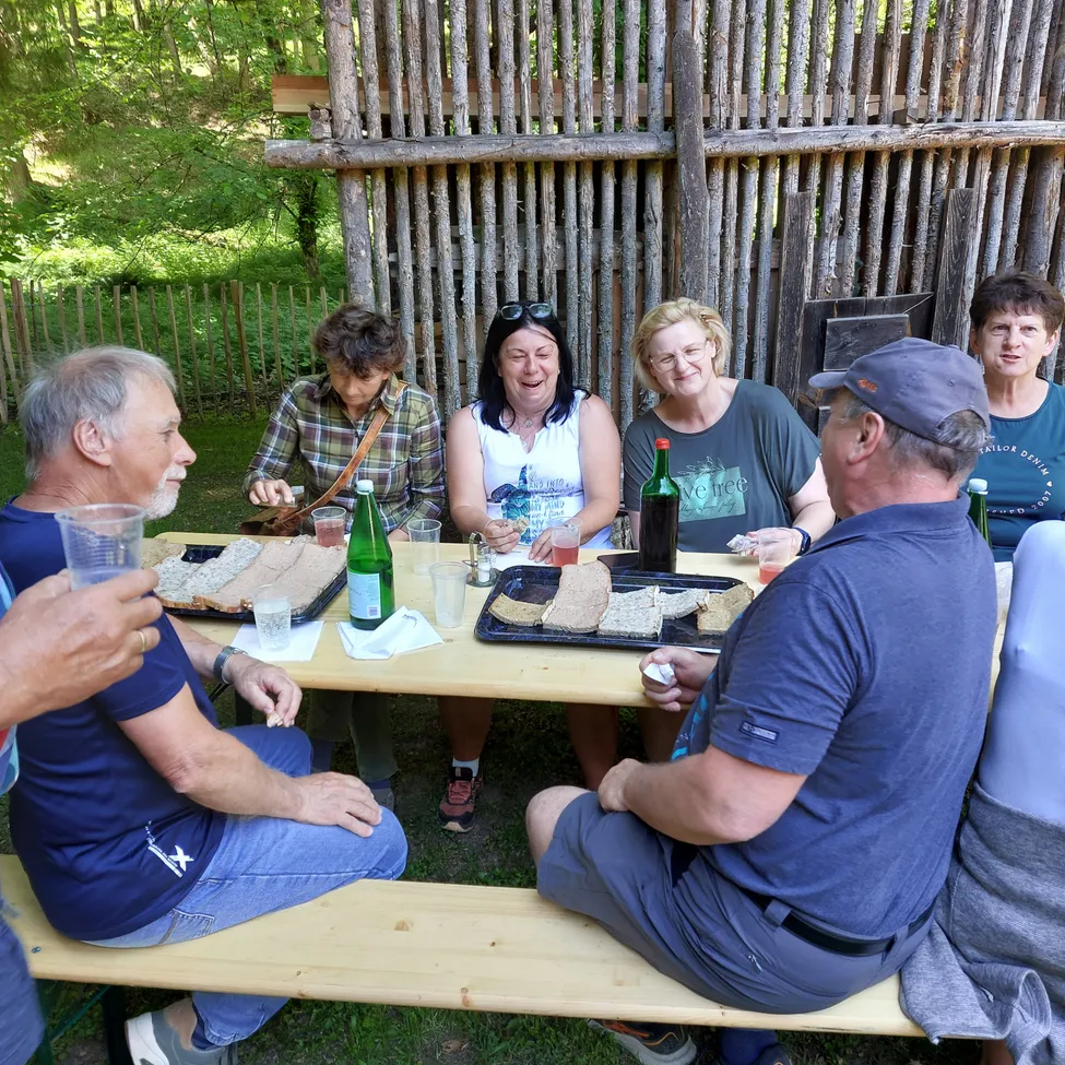Eine Gruppe von Menschen sitzt um einen Holztisch im Freien, lächelt und lacht. Sie genießen Essen und Getränke. Der Tisch ist mit Sandwiches, Getränken und Servietten gedeckt. Hinter ihnen steht ein Holzzaun.