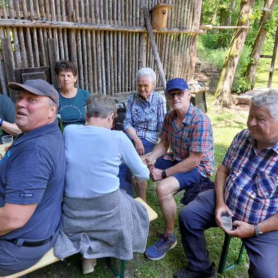 Eine Gruppe älterer Personen, die auf Bänken im Garten sitzen, einer Mann hält ein Glas.