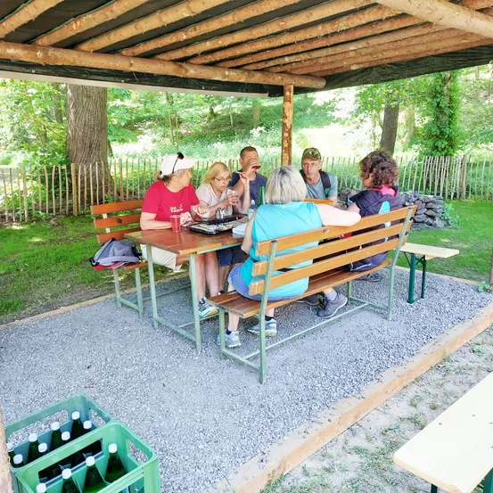 Eine Gruppe von Menschen sitzt um einen Tisch in einem Pavillon und genießt eine Mahlzeit und Getränke. Sie sind von einer Rasenfläche und einem Holzzaun umgeben.