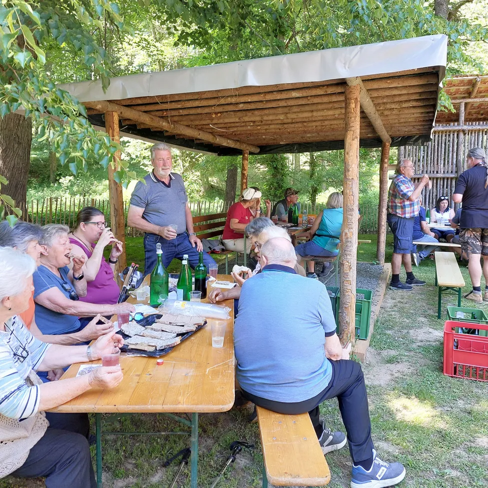 Eine Gruppe von Menschen sitzt unter einem Holzdach in einem Park um einen Tisch herum und genießt Essen und Getränke.