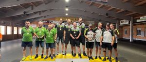 Eine Gruppe von Männern in grünen und schwarzen Shirts und Shorts steht auf einem Podest mit Medaillen um den Hals. Sie befinden sich in einer Sporthalle mit einer Holzdecke und Bannern an der Wand.
