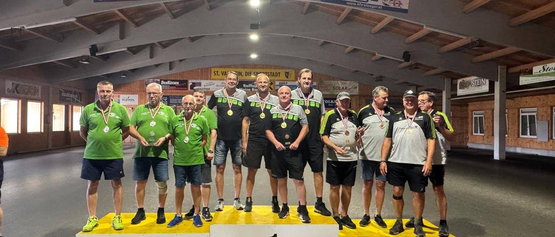Eine Gruppe von Männern in grünen und schwarzen Shirts und Shorts steht auf einem Podest mit Medaillen um den Hals. Sie befinden sich in einer Sporthalle mit einer Holzdecke und Bannern an der Wand.