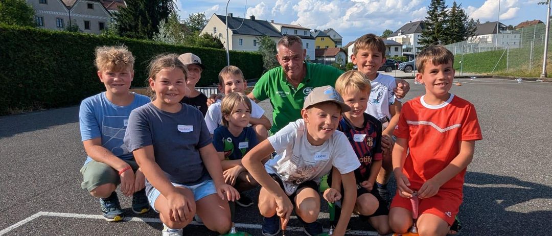 Eine Gruppe von Kindern und einem Mann posiert für ein Foto auf einem Sportplatz mit klarem Himmel und Häusern im Hintergrund.