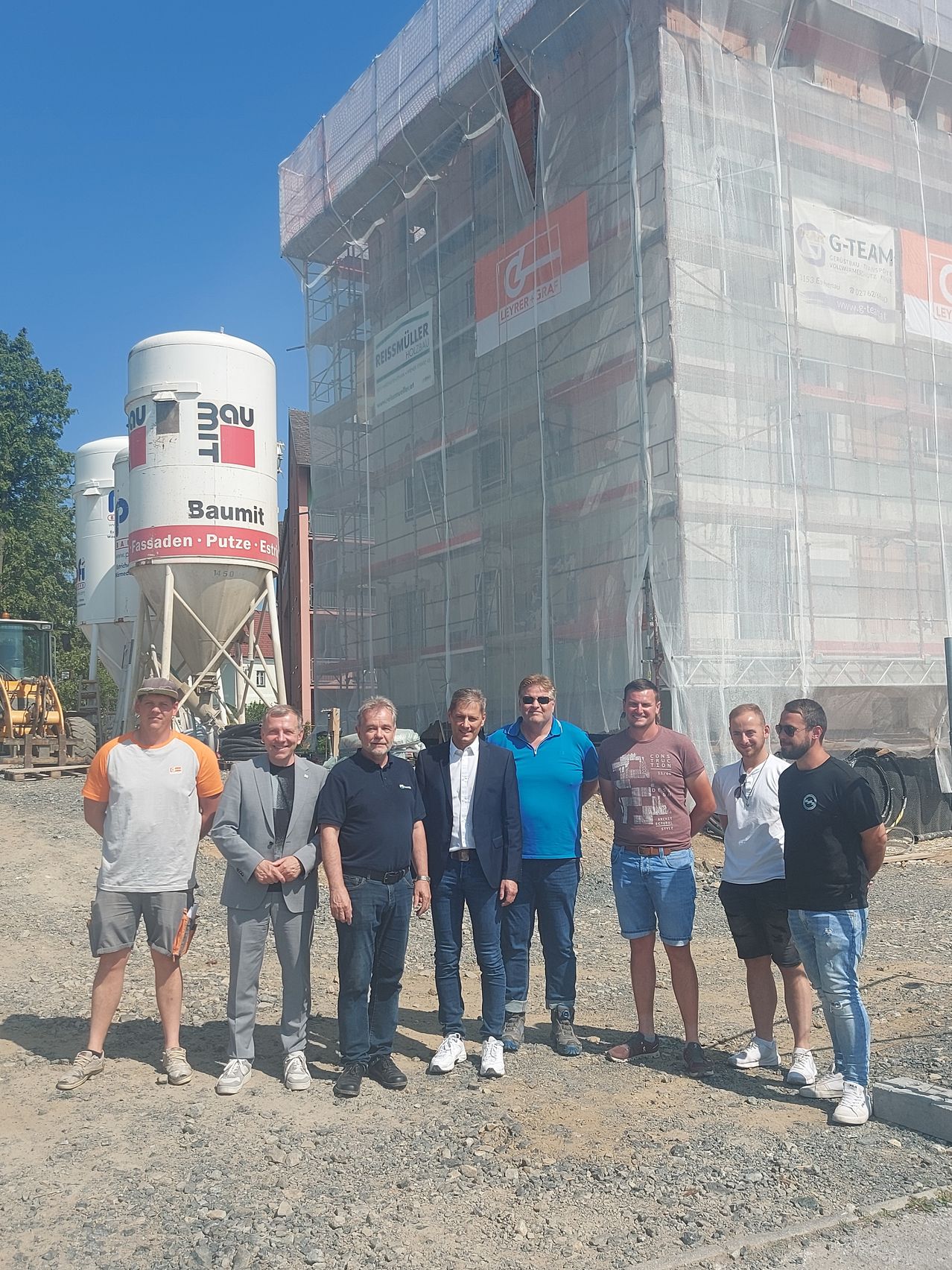 Sieben Männer posieren vor einer Baustelle. Einer der Männer trägt einen Anzug. Hinter ihnen stehen zwei große Lagertanks. Das im Bau befindliche Gebäude hat Glaswände.