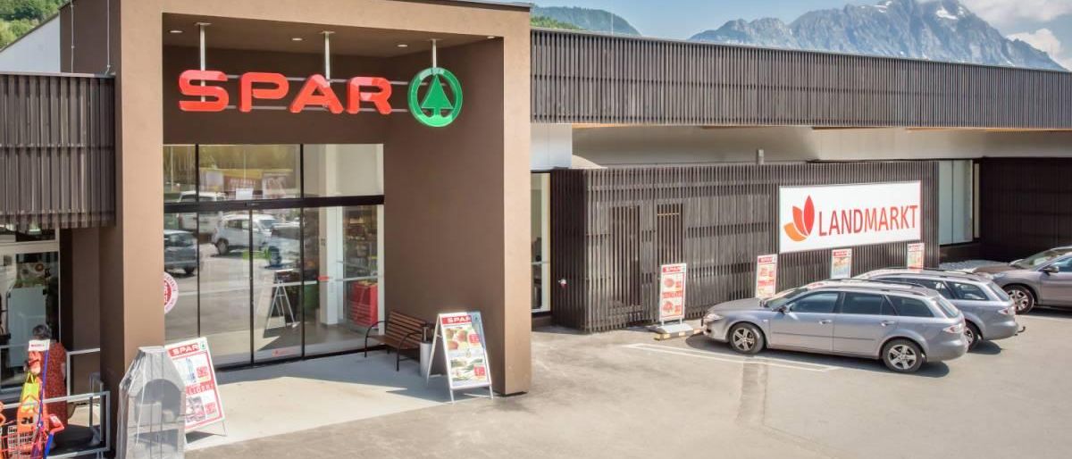 Der Vordereingang eines Spar-Supermarktes. Ein Berg im Hintergrund, Glastüren und geparkte Autos. Eine Bank und Schilder in der Nähe.