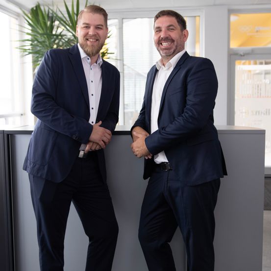 Two men in formal attire standing behind a desk, smiling and looking at the camera.