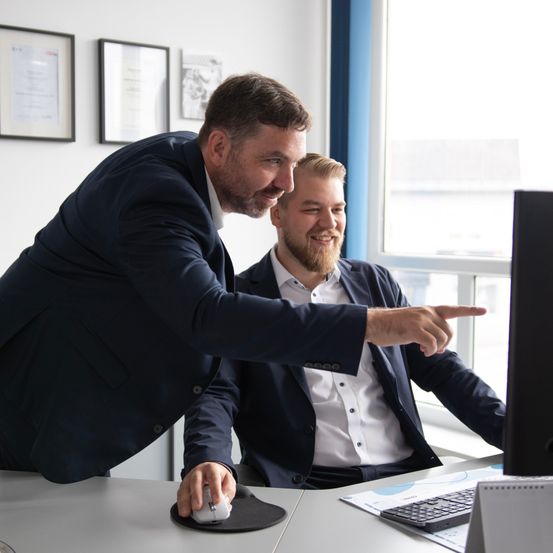 Two men in suits are working together at a desk, one pointing at a computer screen.