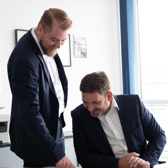Two men in suits are smiling and looking at something on a desk in an office.