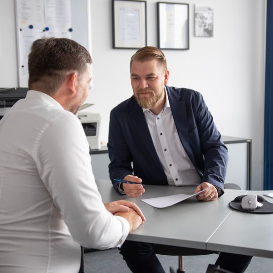 Two men in a professional setting, one in a suit and the other in a white shirt, are engaged in a conversation while sitting at a desk. Papers and a pen are on the desk.