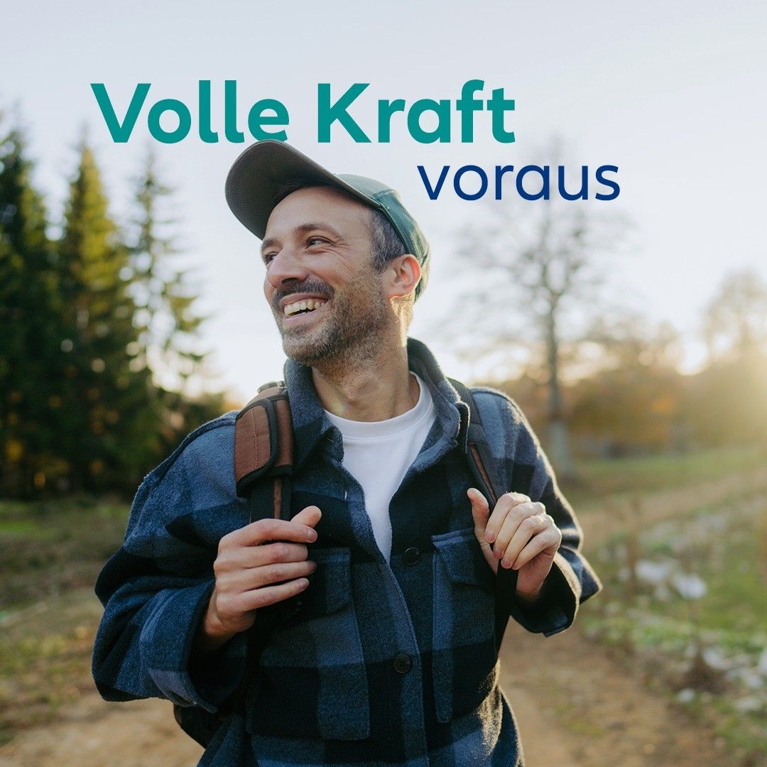 A smiling man with a backpack and a hat is walking on a dirt path. The words 'Volle Kraft voraus' are written above him.