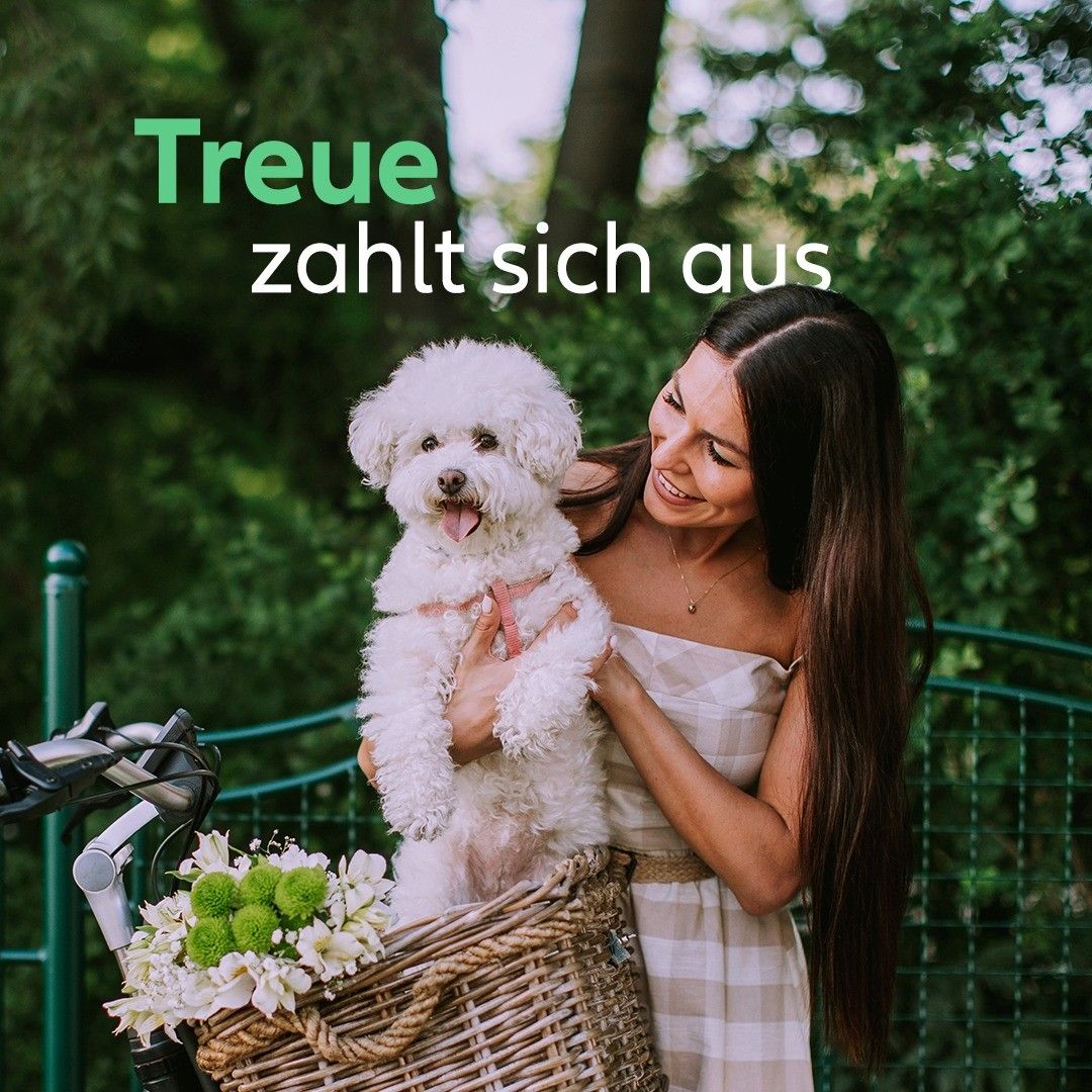 A woman with long hair holds a white dog in a basket on a bicycle. The dog is wearing a pink ribbon. The woman is smiling. The words 'Treue zahlt sich aus' are written on the image.