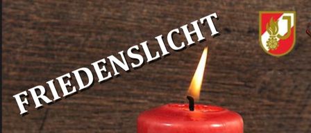 Eine rote Kerze mit dem Wort 'Friedenslicht' darüber brennt mit einer Flamme. Sie hat ein Logo in der Mitte. Ein goldener Stern steht vor der Kerze. Einige Zimtstangen sind auf der rechten Seite der Kerze platziert.