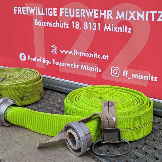 Zwei grüne Feuerwehrschläuche liegen vor einem roten Banner. Das Banner trägt die Aufschrift 'Freiwillige Feuerwehr Mixnitz' und 'Barenschutz 18, 8131 Mixnitz'. Es sind auch Social-Media-Icons zu sehen.