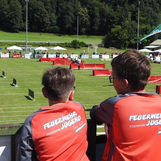 Zwei Jungen in roten und grauen Trikots beobachten eine Sportveranstaltung. Sie stehen am Rand eines Feldes mit roten Barrieren. Im Hintergrund sind Menschen unter Schirmen versammelt, und das Feld hat Tore.