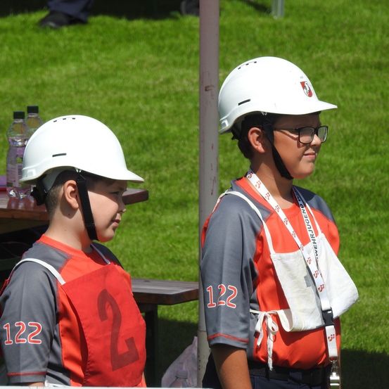 Zwei Personen in orangefarbenen Westen und weißen Helmen stehen auf einem Rasenfeld. Ein Tisch mit Wasserflaschen ist in der Nähe. Eine Person hat die Nummer 2 auf ihrer Weste, die andere hat die Nummer 122.