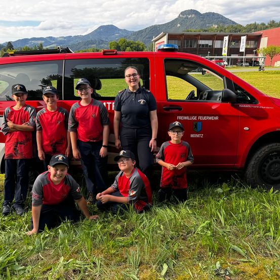 Eine Gruppe junger Kinder in roten und grauen Uniformen posiert vor einem roten Fahrzeug mit einer Frau in Uniform.