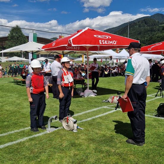 Zwei Kinder in roten Westen stehen auf einem Feld, ein Mann in Uniform neben ihnen. Sie stehen unter einem roten Zelt. Im Hintergrund sitzen Menschen auf Bänken.