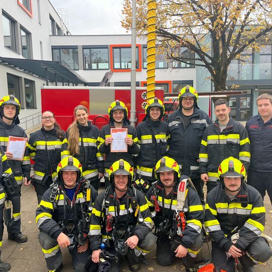 Eine Gruppe Feuerwehrleute in voller Ausrüstung posiert für ein Foto vor einer Feuerwache mit einem roten Feuerwehrauto im Hintergrund.