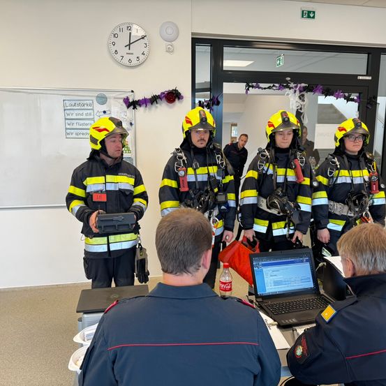 Eine Gruppe Feuerwehrleute in voller Ausrüstung steht in einem Raum mit einer Uhr und einer Tafel. Ein Laptop, eine Flasche und Taschen stehen auf einem Tisch vor ihnen.