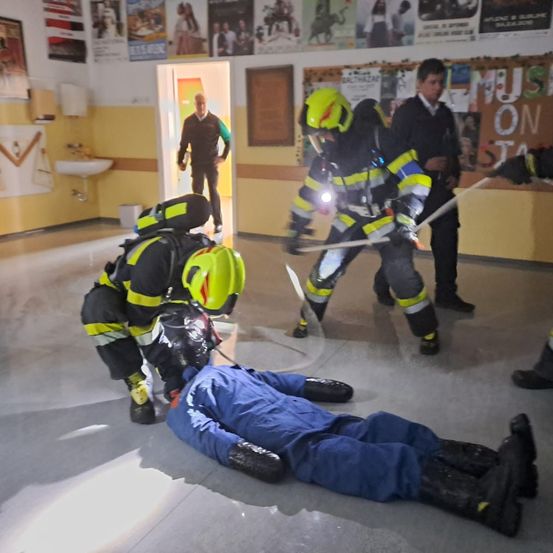 Zwei Feuerwehrleute üben Rettungsmanöver mit einem Mann, der auf dem Boden liegt. Sie tragen vollständige Schutzausrüstung, einschließlich Helme und Stiefel. Eine weitere Person beobachtet von der Tür aus.
