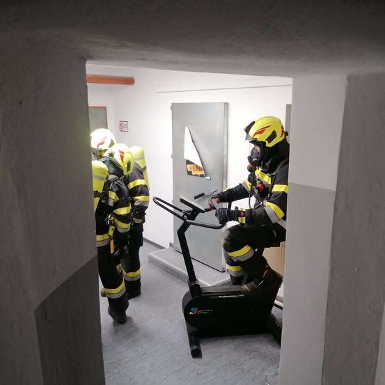 Drei Feuerwehrleute in gelben und schwarzen Uniformen, einer mit Gasmaske, sind in einem Raum. Der Mann auf dem Fahrrad trägt einen gelben Helm.