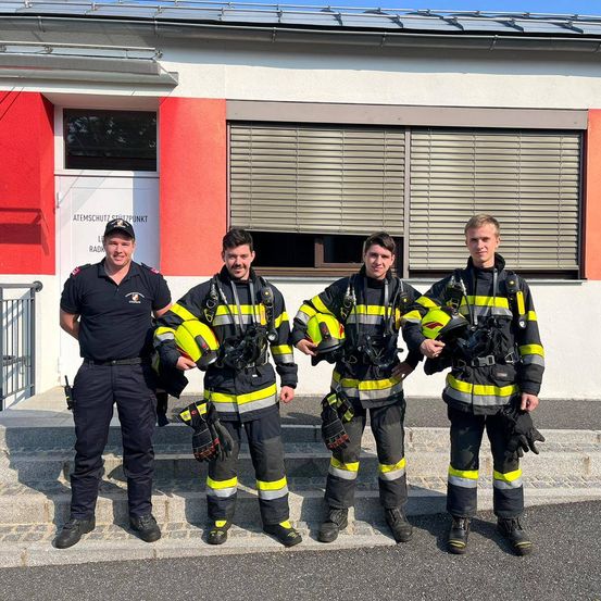 Vier Feuerwehrleute in gelben und schwarzen Uniformen stehen vor einem Gebäude mit rot-weißen Wänden. Einer hält einen Helm, und sie tragen alle Handschuhe.