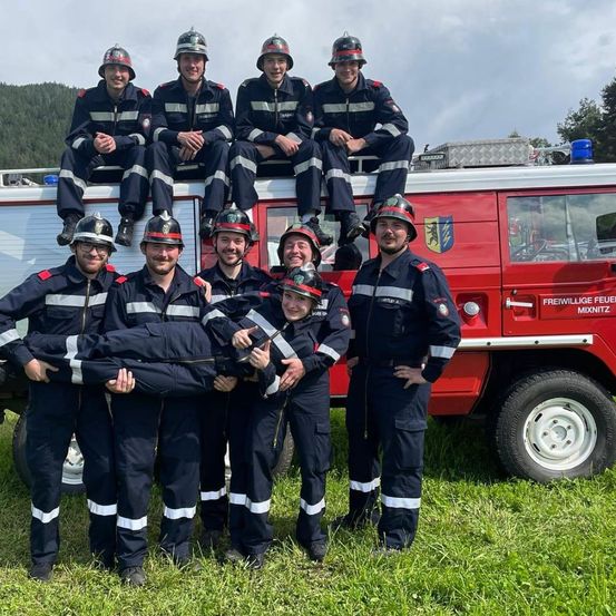 Eine Gruppe von Feuerwehrleuten in Uniformen und Helmen posiert für ein Foto. Sie stehen vor einem roten Feuerwehrwagen. Einige sitzen auf dem Wagen, während andere auf dem Boden stehen.