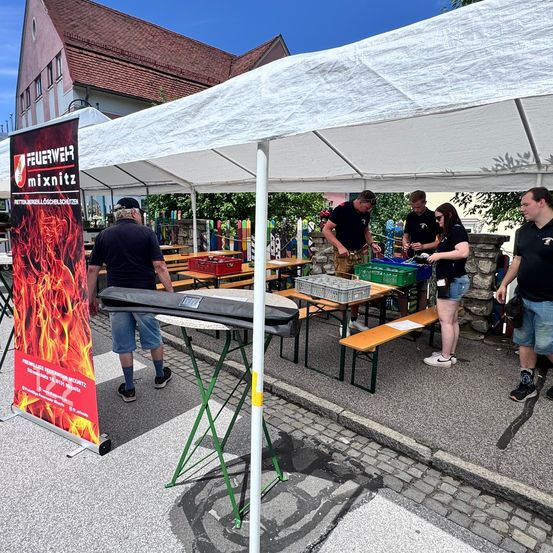 Feuerwehrleute richten einen Vordach, Tische und Bänke im Freien auf. Eine Gruppe von Menschen ordnet Gegenstände auf den Tischen an. Ein roter Banner mit weißem Text steht in der Nähe.