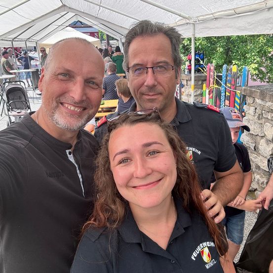 Drei Personen in schwarzen Uniformen lächeln für ein Selfie bei einer Outdoor-Veranstaltung. Hinter ihnen ist ein weißes Zelt und eine Steinmauer zu sehen, mit mehreren Personen und Tischen im Hintergrund.