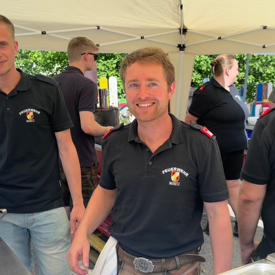 Eine Gruppe von Menschen in Uniform, wahrscheinlich Feuerwehrleute, steht in einer Outdoor-Umgebung unter einem Zelt. Ein Mann lächelt und trägt einen Gürtel.