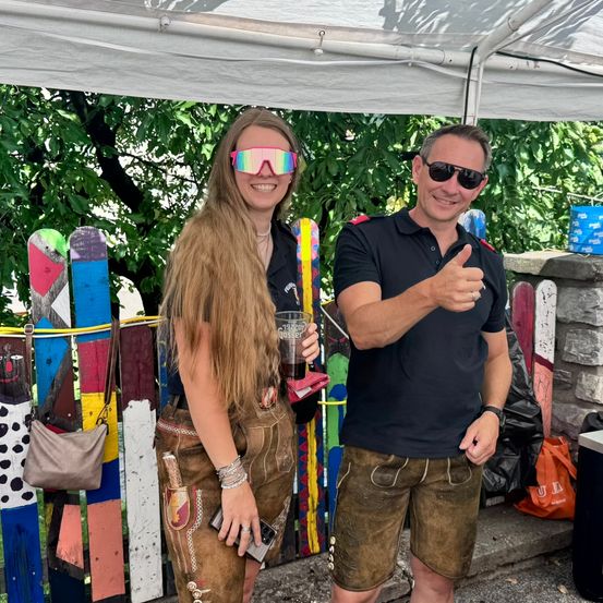 Eine Frau in traditioneller bayerischer Kleidung und ein Mann in einem schwarzen Hemd lächeln für die Kamera draußen unter einem Zelt. Sie stehen neben einem farbenfrohen Zaun mit Bäumen im Hintergrund.