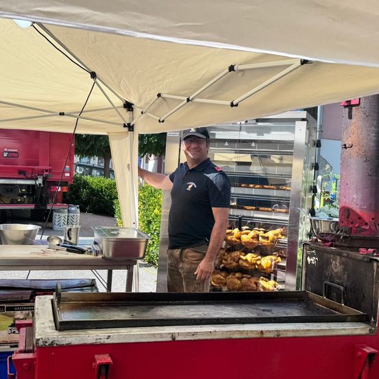 Ein Mann steht unter einem Zelt und lächelt in die Kamera. Er bereitet Essen vor einem offenen Grill zu. Mehrere Tabletts mit Essen sind hinter ihm ausgestellt.