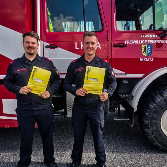 Zwei Feuerwehrleute stehen vor einem Feuerwehrauto und halten gelbe Bücher. Das Auto ist mit 'Freiwillige Feuerwehr' und einem Logo gekennzeichnet. Beide Männer tragen Uniformen und einer hält ein Buch mit dem Titel 'Fahrtechnik'.