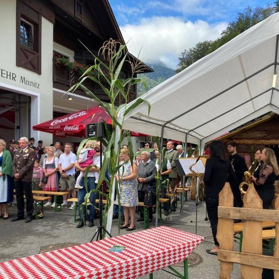 Eine Gruppe von Menschen ist im Freien versammelt, einige stehen und andere sitzen. In der Mitte steht ein Maiskolben. Musikinstrumente, einschließlich Saxophone, sind vorhanden. Hinter der Gruppe befindet sich ein Gebäude mit einem Coca-Cola-Schild.