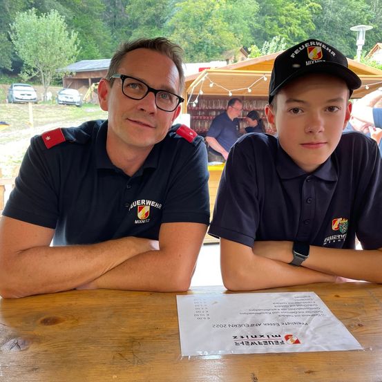 Zwei Männer, beide in schwarzen Uniformen, sitzen an einem Tisch. Ein Mann trägt eine Mütze mit der Aufschrift 'FEUERWEHR'. Sie sitzen neben einem Stück Papier. Bäume und ein Gebäude im Hintergrund.