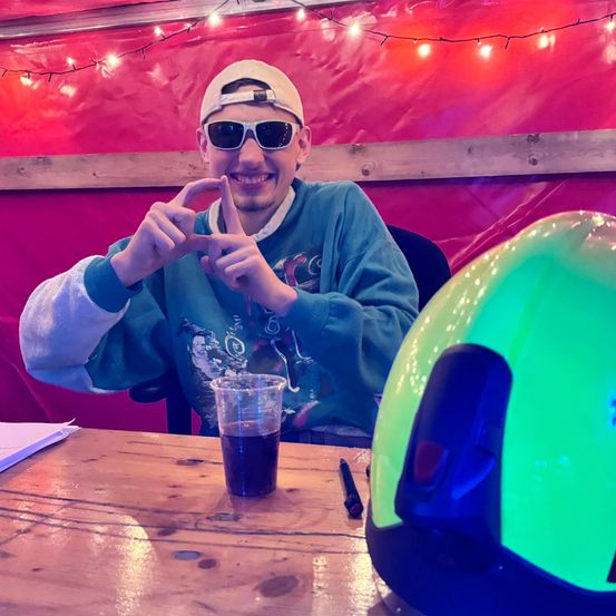 Ein Mann mit Cap und Sonnenbrille sitzt an einem Tisch mit einem Glas dunkler Flüssigkeit, lächelt und macht ein Friedenszeichen. Daneben steht ein grüner Helm. Dahinter ist ein roter Hintergrund mit Lichtern.