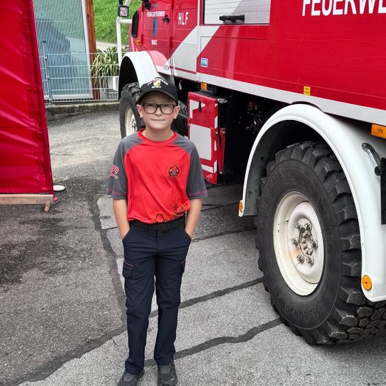 Ein Junge steht vor einem roten Feuerwehrauto mit dem Wort 'Feuerwehr' darauf. Er trägt ein rotes Shirt mit Logo und schwarze Hosen.