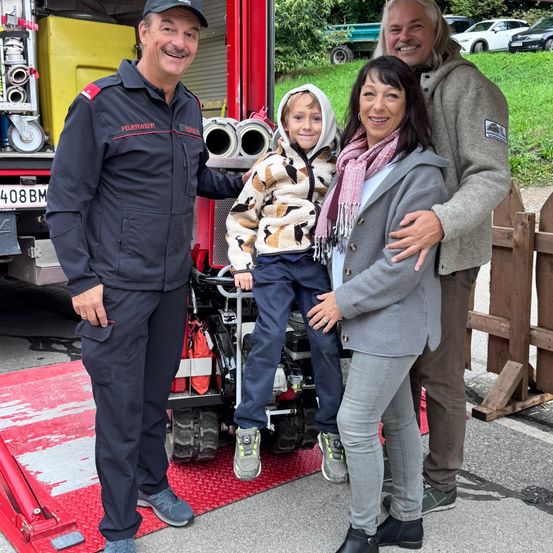 Eine vierköpfige Familie steht vor einem Feuerwehrauto. Der Mann in Uniform lächelt, und ein Kind sitzt auf dem Auto. Die Frau trägt einen Schal, und sie alle posieren für ein Foto.