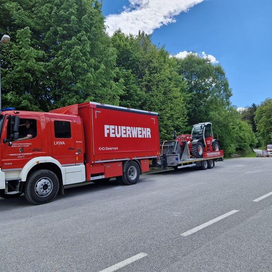 Ein roter Feuerwehrwagen zieht einen roten Traktor hinter sich her und fährt eine Straße hinunter. Der Himmel ist klar mit einigen Wolken. Bäume säumen beide Seiten der Straße.