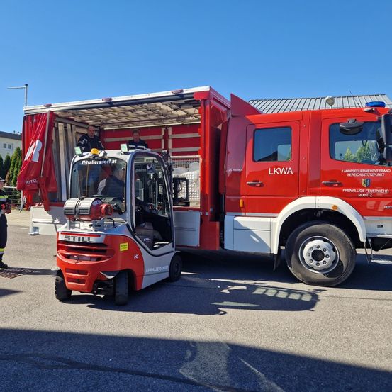 Ein roter Feuerwehrwagen steht mit geöffneter Hecktür. Im Inneren fährt eine Person einen Gabelstapler, und zwei weitere stehen im Inneren. Der Wagen ist mit 'LKWA' und 'Feuerwehr' gekennzeichnet.