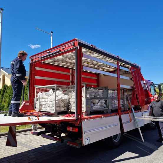 Ein roter LKW mit offener Ladefläche ist auf einer Straße geparkt. Im Inneren des LKW befinden sich zwei Metallkisten mit weißen Säcken. Zwei Männer stehen neben dem LKW. Einer steht auf der Plattform des LKW und der andere daneben.