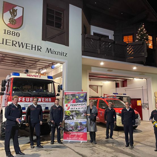 Eine Gruppe von Feuerwehrleuten steht vor einer Feuerwehrstation namens Feuerwehr Mixnitz. Sie tragen Uniformen und stehen in der Nähe eines Feuerwehrwagens.
