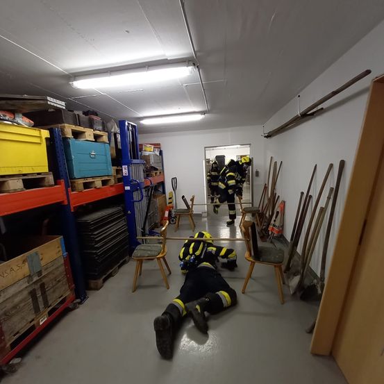 Zwei Feuerwehrleute in Schutzkleidung trainieren in einem Lagerraum mit gestapelten Holzkisten und verschiedenen Werkzeugen an der Wand. Ein Feuerwehrmann liegt auf dem Boden.