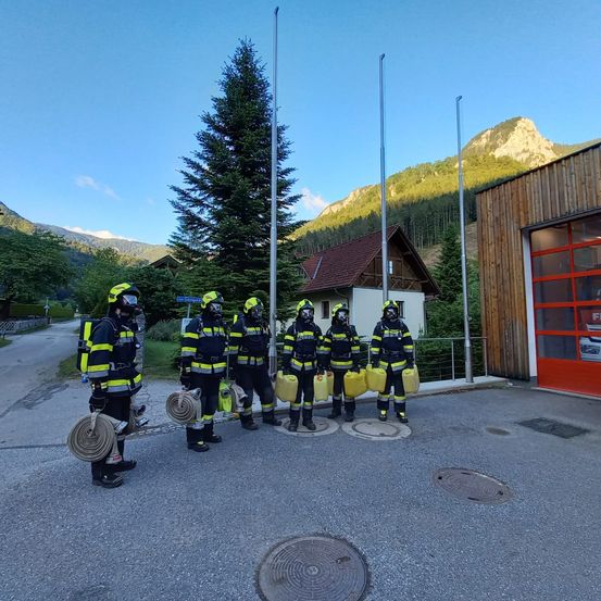 Eine Gruppe von Feuerwehrleuten in voller Ausrüstung steht in der Nähe einer Feuerwache, mit Bergen im Hintergrund.