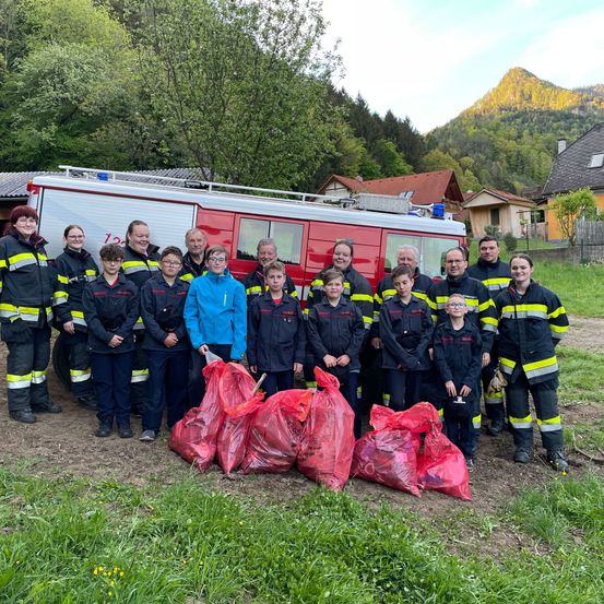 Eine Gruppe von Feuerwehrleuten und Kindern posiert für ein Foto mit einem Feuerwehrwagen und Säcken voller Müll. Sie stehen in einem Grasbereich mit Häusern und einem Berg im Hintergrund.