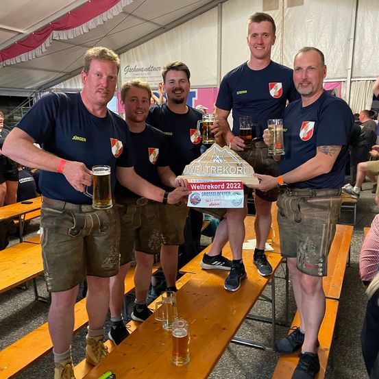 Fünf Männer stehen auf einer Bank und halten eine Trophäe und Gläser Bier. Sie tragen passende blaue T-Shirts und grüne Shorts. Ein Mann hält ein Schild mit den Worten 'Weltrekord 2022'.