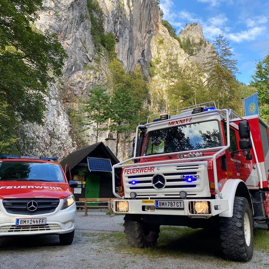 Zwei Feuerwehrfahrzeuge sind auf einer Kiesfläche mit einem Berg im Hintergrund geparkt. Das vordere Feuerwehrfahrzeug hat das Wort 'Feuerwehr' auf seiner Front. Dahinter steht ein kleines Gebäude mit einem Solarpanel.