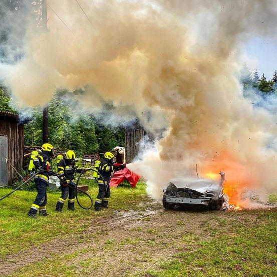 Feuerwehrleute löschen ein brennendes Auto in einer ländlichen Umgebung, wobei Rauch aufsteigt.