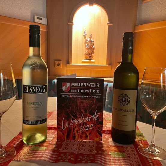 Zwei Weinflaschen, eine mit der Aufschrift Elsneg und die andere mit Frauwallner, stehen neben einem Buch mit dem Titel 'Weihnachtskarte 2022' auf einem Tisch. Zwei Weingläser sind ebenfalls vorhanden. Im Hintergrund ist eine kleine Statue zu sehen.