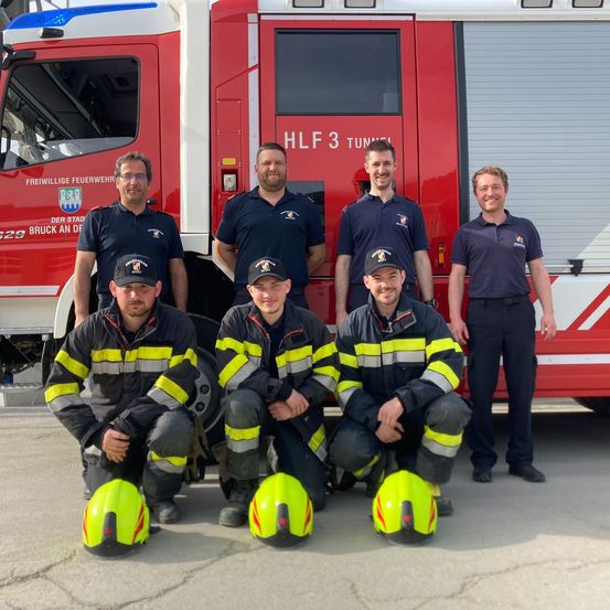 Sechs Feuerwehrleute posieren vor einem roten Feuerwehrwagen. Der Wagen hat die Aufschrift 'HLF 3' und 'TUNEL'. Alle tragen Uniformen und Helme.